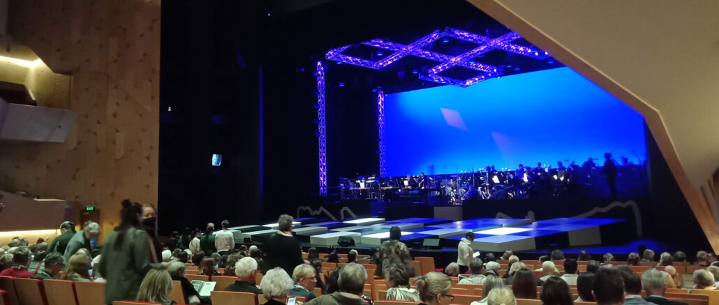A view of the large auditorium of the Kiri Te Kanawa Theatre. There are rows of audience members facing the stage, while others are taking their seats. Few people appear to be wearing masks.