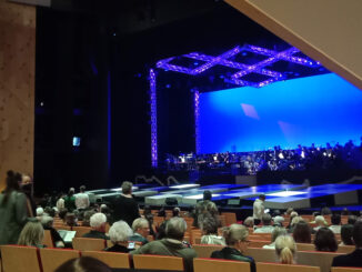 A view of the large auditorium of the Kiri Te Kanawa Theatre. There are rows of audience members facing the stage, while others are taking their seats. Few people appear to be wearing masks.